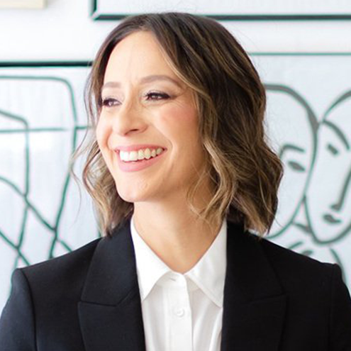 A headshot of Olga Brandeis sitting in a director's chair in front of a gallery wall with black and white, framed, drawings.